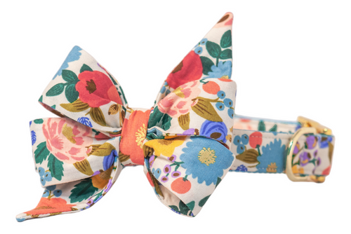 Floral dog collar and belle bow set crafted from a soft cream base with an intricate garden print of bold, multicolored flowers and berries. The oversized belle bow, shaped with two upward flared points and two downward tails, adds a whimsical and feminine flair. A shiny buckle and loop accent the collar’s structure.