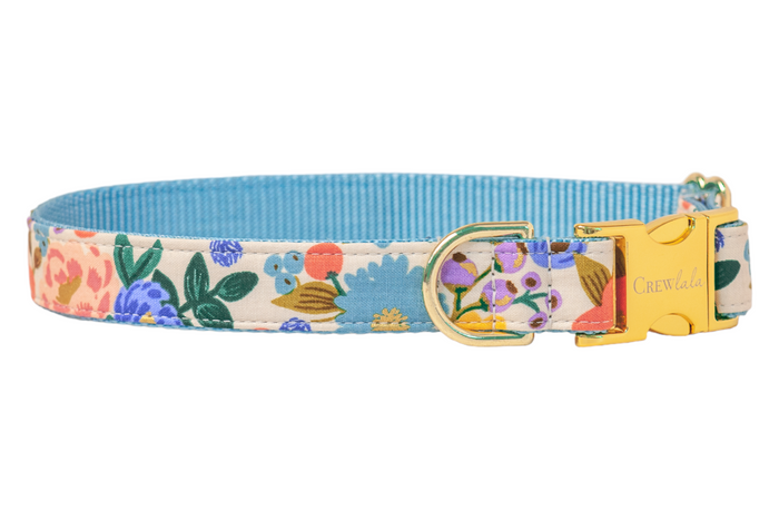Dog collar with a colorful floral pattern on a cream background, featuring flowers with leaves and metallic detailing. The collar has a vibrant nylon underside and metal hardware, including a side-release buckle engraved with “Crewlala” and a D-ring.