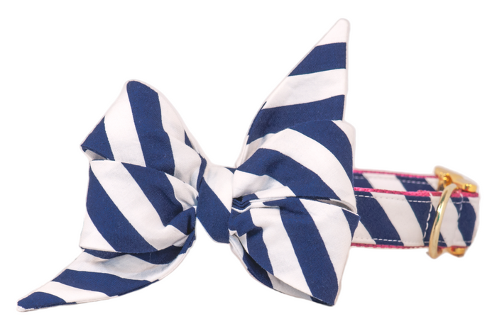 A cheerful white golden retriever sits on a sidewalk wearing a crisp navy blue and white striped bow tie, perfectly coordinated with a matching leash held by a person in a white dress. The pup’s tongue hangs out in a happy pant, radiating charm and nautical flair.