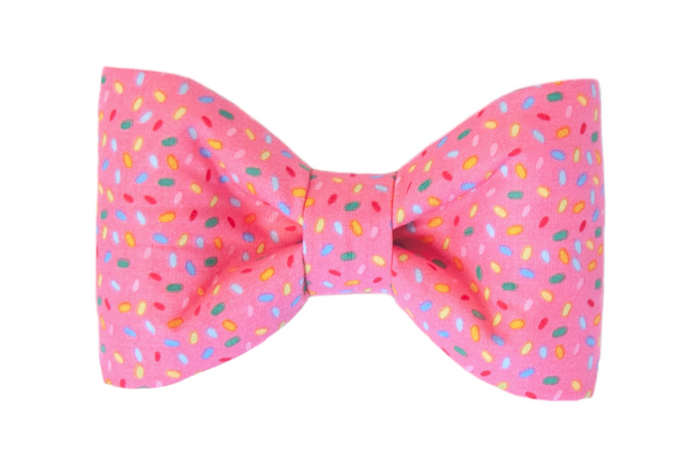 Playful dog bow tie made from bright pink fabric covered in multicolored sprinkle shapes. The bow has a classic shape with softly curved sides.