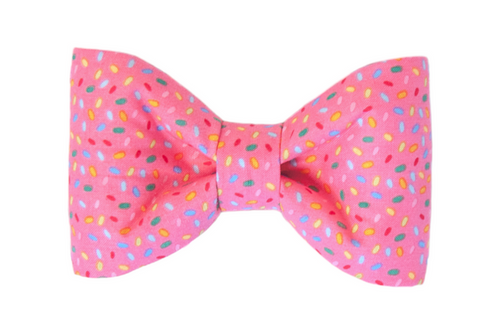 Playful dog bow tie made from bright pink fabric covered in multicolored sprinkle shapes. The bow has a classic shape with softly curved sides.