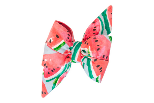 A large, decorative feminine dog belle bow made from fabric printed with a vibrant watermelon design. The pattern features pink-red watermelon slices with black seeds, white rinds, and green outer stripes on a pale blue background. The bow has wide, full loops and pointed tails for a playful, summery look.